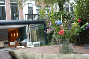 A patio with a glass vase of flowers, a seating area with a table and chairs, and a building with large windows and a balcony in the background.