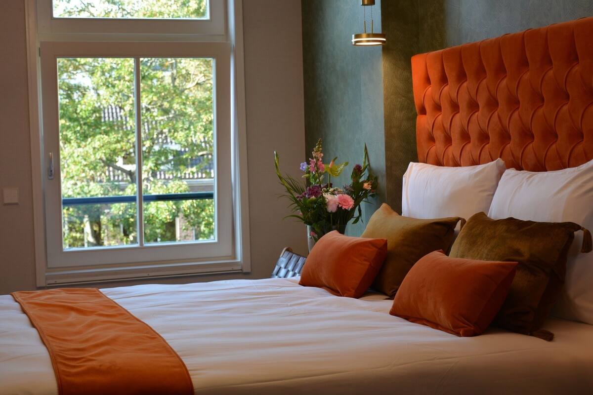 A neatly made bed with a white and orange bedspread, a tufted orange headboard, and a vase with flowers on a nightstand.