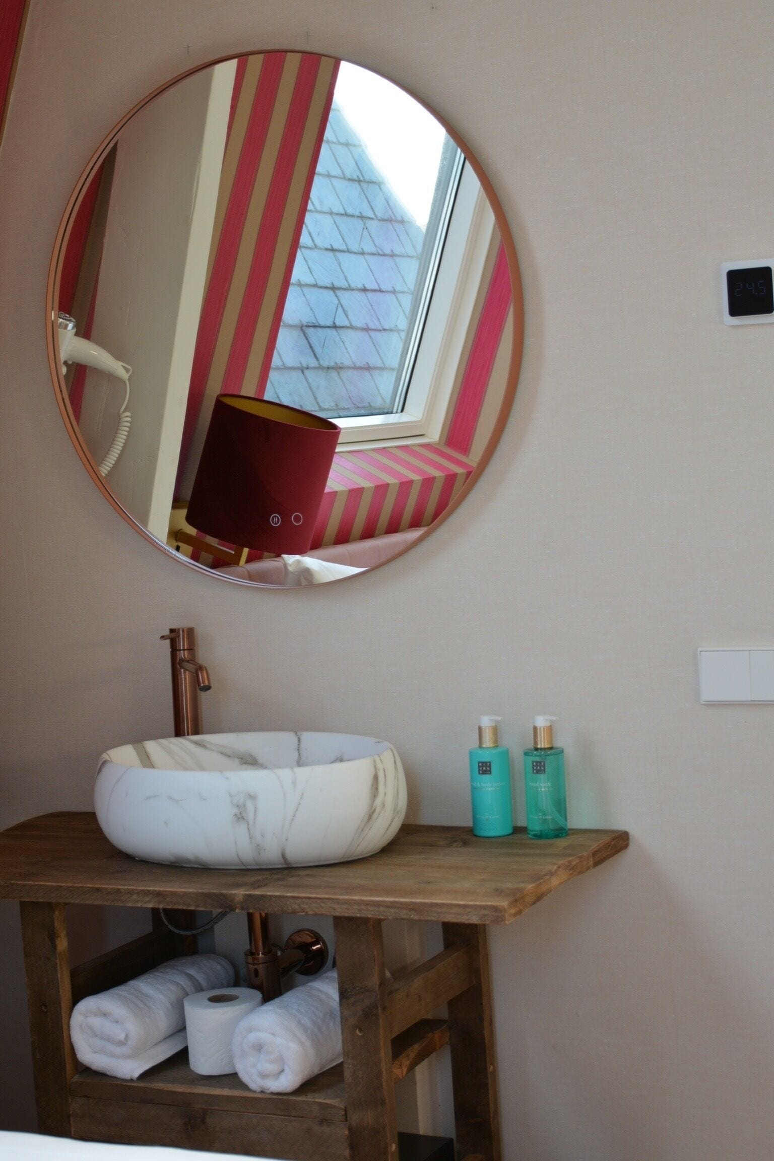 A bathroom with a round mirror, a sink, and a wooden shelf with towels and toiletries.