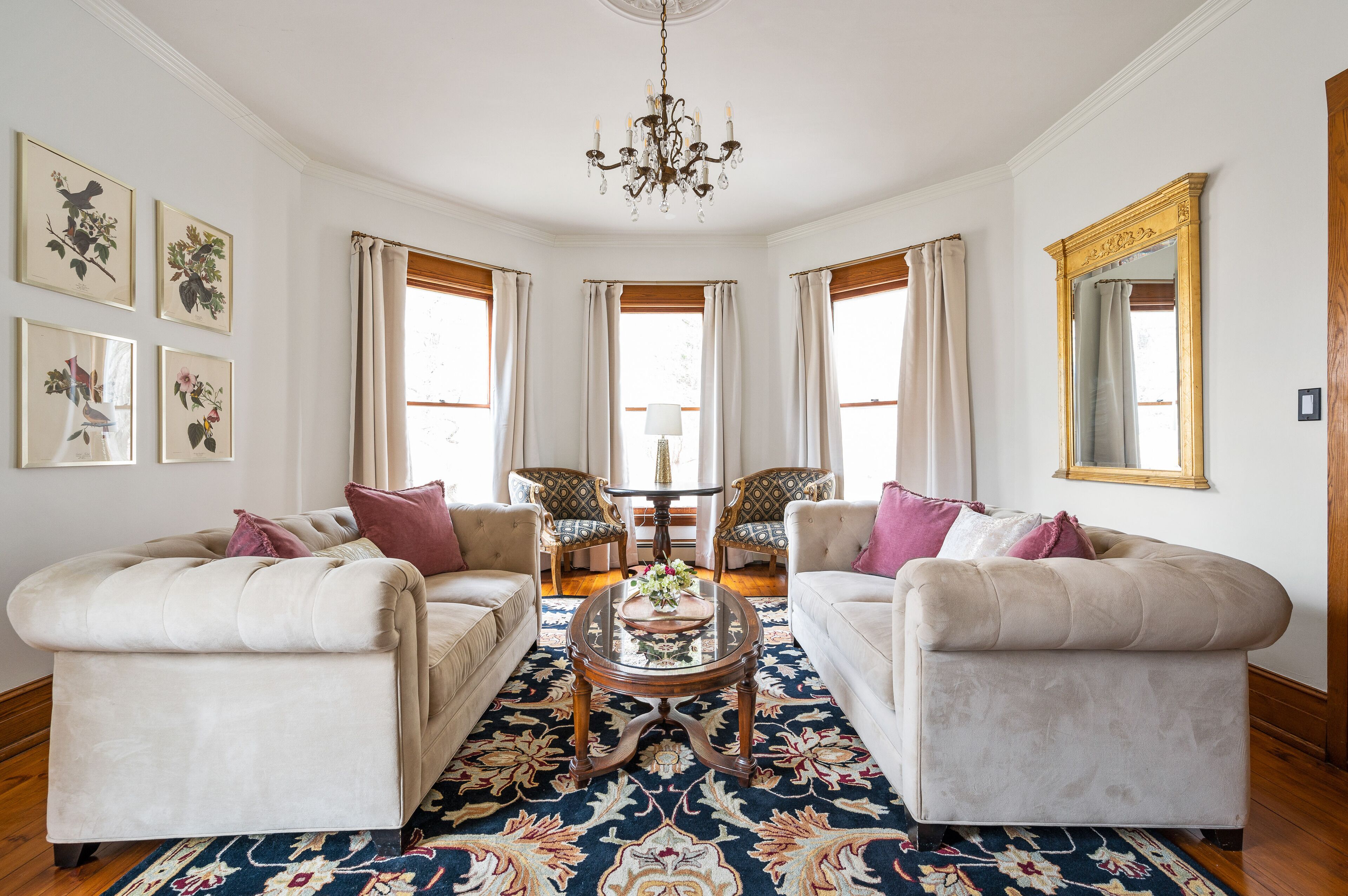 A room with two sofas, a coffee table, and a chandelier. There are framed botanical prints on the wall and a large mirror.