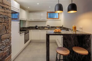 Una cocina moderna con armarios blancos, un muro decorativo de piedra, encimera de mármol y dos taburetes de madera.