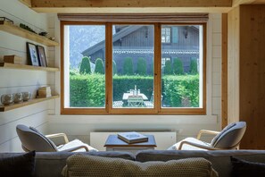 Une pièce chaleureuse avec une grande fenêtre, un canapé, deux fauteuils, une petite table et des étagères en bois.