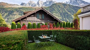 Um chalé de madeira com área de refeições, incluindo uma mesa e cadeiras, cercado por sebes bem cuidadas e uma paisagem montanhosa ao fundo.