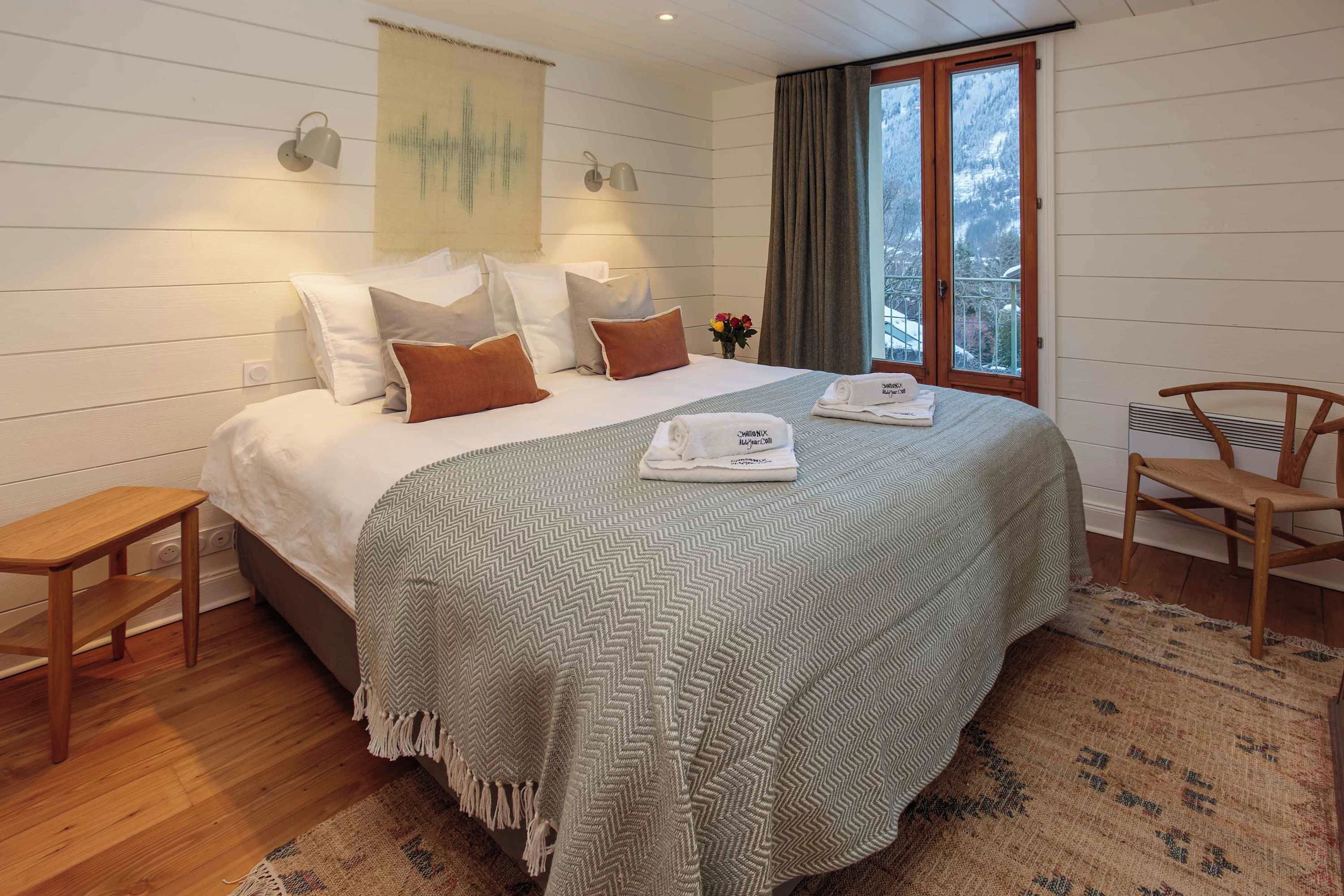 A neatly made bed with a patterned bedspread, two bedside lamps, a small table, a chair, and a window with curtains.