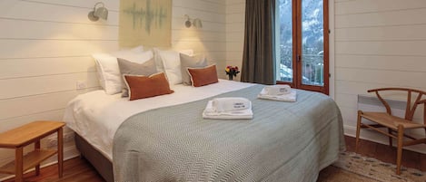 A neatly made bed with a patterned bedspread, two bedside lamps, a small table, a chair, and a window with curtains.