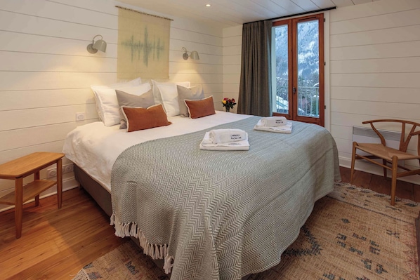 A neatly made bed with a patterned bedspread, two bedside lamps, a small table, a chair, and a window with curtains.