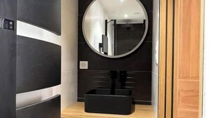 Une petite salle de bain avec un mur recouvert de carreaux noirs et blancs, un miroir rond, un lavabo noir et un meuble-lavabo en bois doté de tiroirs.