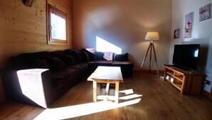 Una sala de estar con piso de madera, un sofá marrón, una mesita de madera, una lámpara de pie y un televisor sobre un mueble de madera.