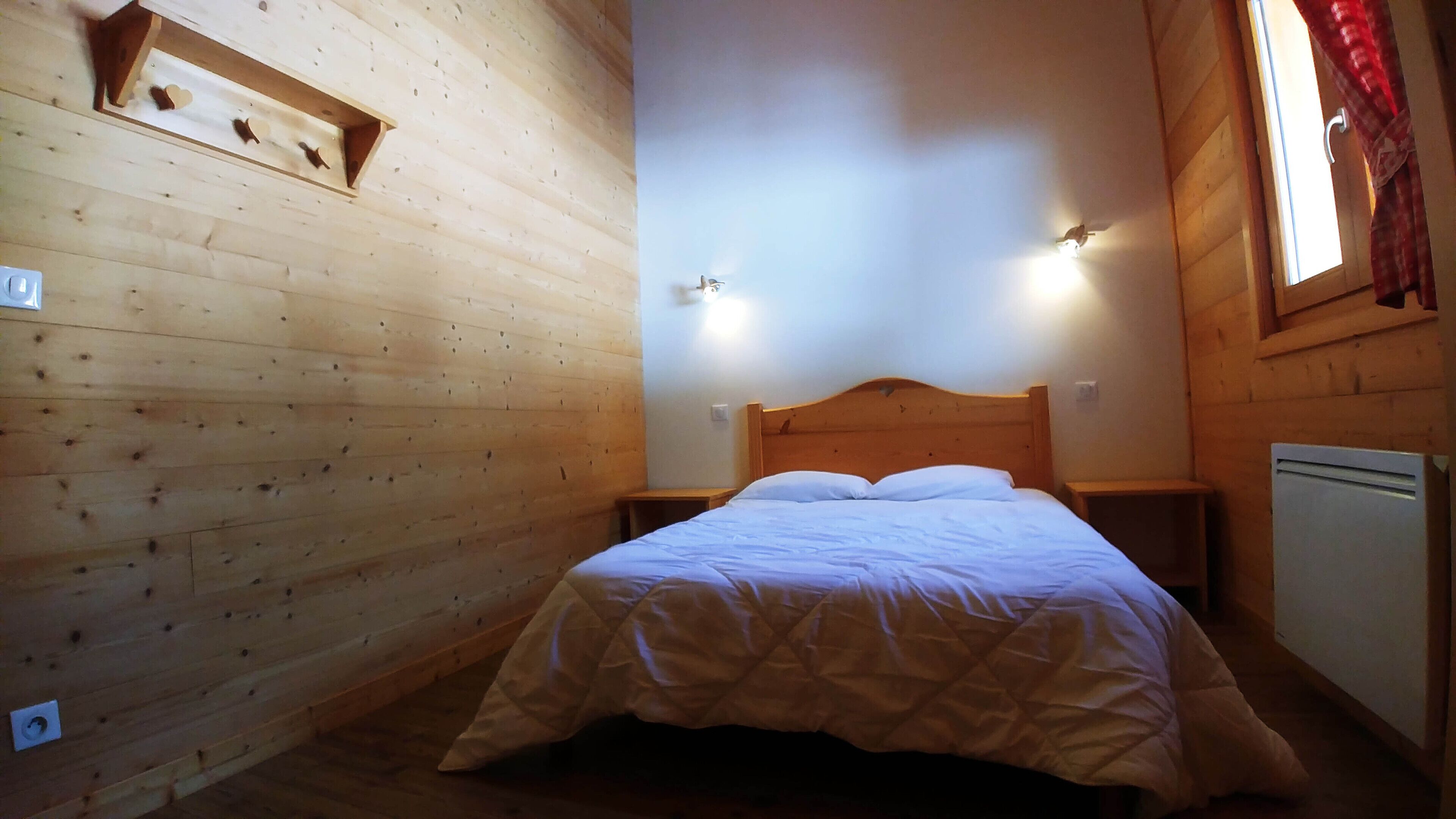 A wooden cabin room with a bed, two bedside tables, a wall-mounted shelf, and a window with curtains.