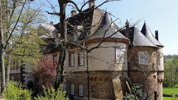 Un grand bâtiment historique en pierre, doté d’une tour ronde, entouré d’une végétation luxuriante et surplombé d’un ciel dégagé.