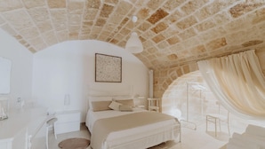 A bedroom with a stone ceiling, a bed with white bedding, a bedside table, and a large window with curtains. - Casa Vacanze Portanuova Monopoli (Monopoli)