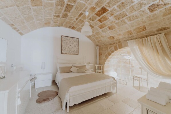 A bedroom with a stone ceiling, a bed with white bedding, a bedside table, and a large window with curtains.