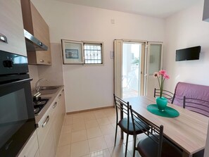 Uma cozinha compacta com fogão, pia e armários. Há uma mesa de jantar com cadeiras e um pequeno sofá. Uma TV está fixada na parede.