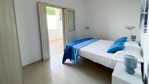 A bedroom with a bed, a nightstand, a lamp, and a window with a view of the outdoors. - Appartamento 69 in Casalabate (Squinzano)