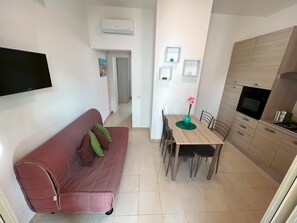 Uma sala de estar com sofá vermelho, mesa de jantar com cadeiras, área de cozinha com armários e fogão, e uma TV de tela plana fixada na parede.