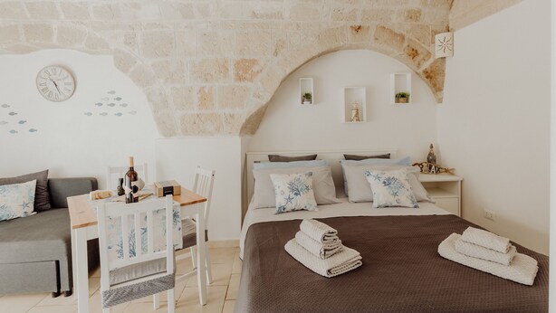 Une pièce chaleureuse avec un mur en pierre, un lit recouvert d’une couverture marron, une chaise en bois, une petite table avec une bouteille et une horloge au mur.