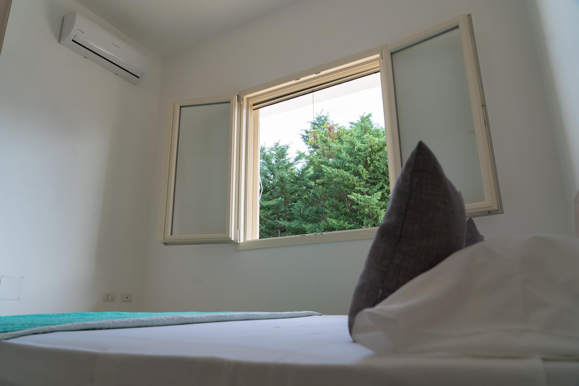 Een slaapkamer met een bed, een raam met uitzicht op bomen, en een airconditioner aan de muur.