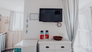 A hotel room with a flat-screen TV mounted on the wall, a white bedside table with a basket, and a window with curtains. - Casa Azzurra in Capitolo (Monopoli)