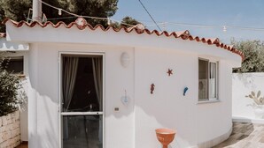 A small, single-story building with a white exterior and terracotta roof tiles. It features a large window with a curtain, a smaller window, and a wall-mounted light fixture. Decorative elements include a heart-shaped ornament and a small star. - Casa Azzurra in Capitolo (Monopoli)