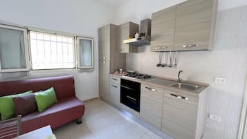Cozinha compacta com eletrodomésticos modernos, forno embutido e pia. Na área de estar adjacente, há um sofá vermelho com almofadas verdes.