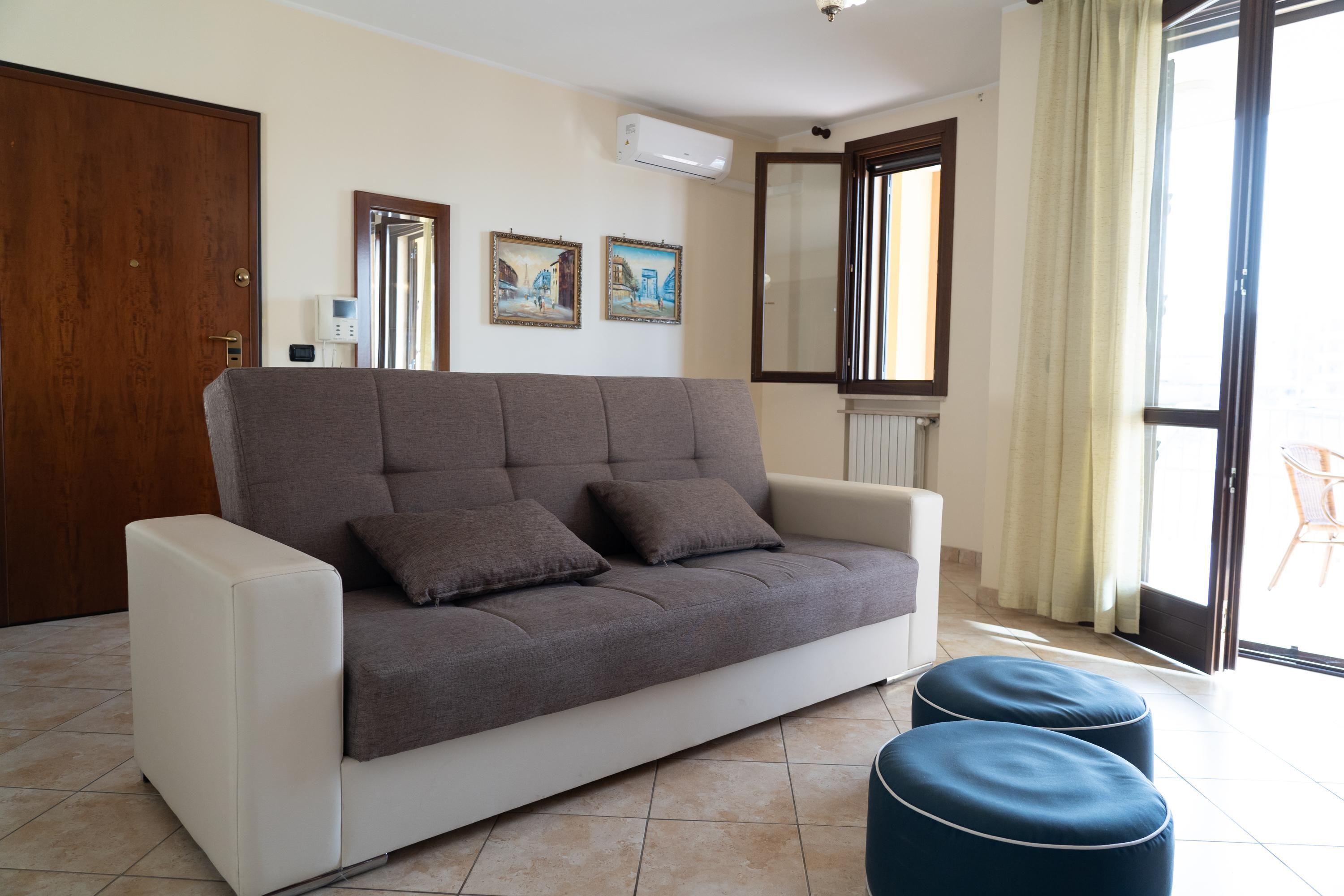 A living room with a sofa, two ottomans, a wooden door, and a window with curtains.