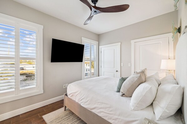 A bedroom with a bed, a ceiling fan, a TV mounted on the wall, and a window with blinds.