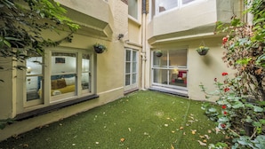 Une cour avec une pelouse artificielle, des jardinières suspendues et une vue sur une pièce comprenant un lit et une fenêtre.
