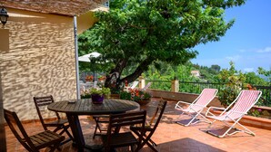An outdoor patio with a round wooden table and chairs, surrounded by potted plants and a large tree providing shade. - Il Ciliegio ,'le Calme et la Tranquillité Sont Notre Carte de Visite' (Castelbuono)