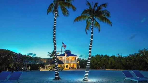 A beachfront hotel with two palm trees, a flag, and a pool area with lounge chairs. - Florida Keys Getaway - Slps 4 -  Ocean View (Tavernier)