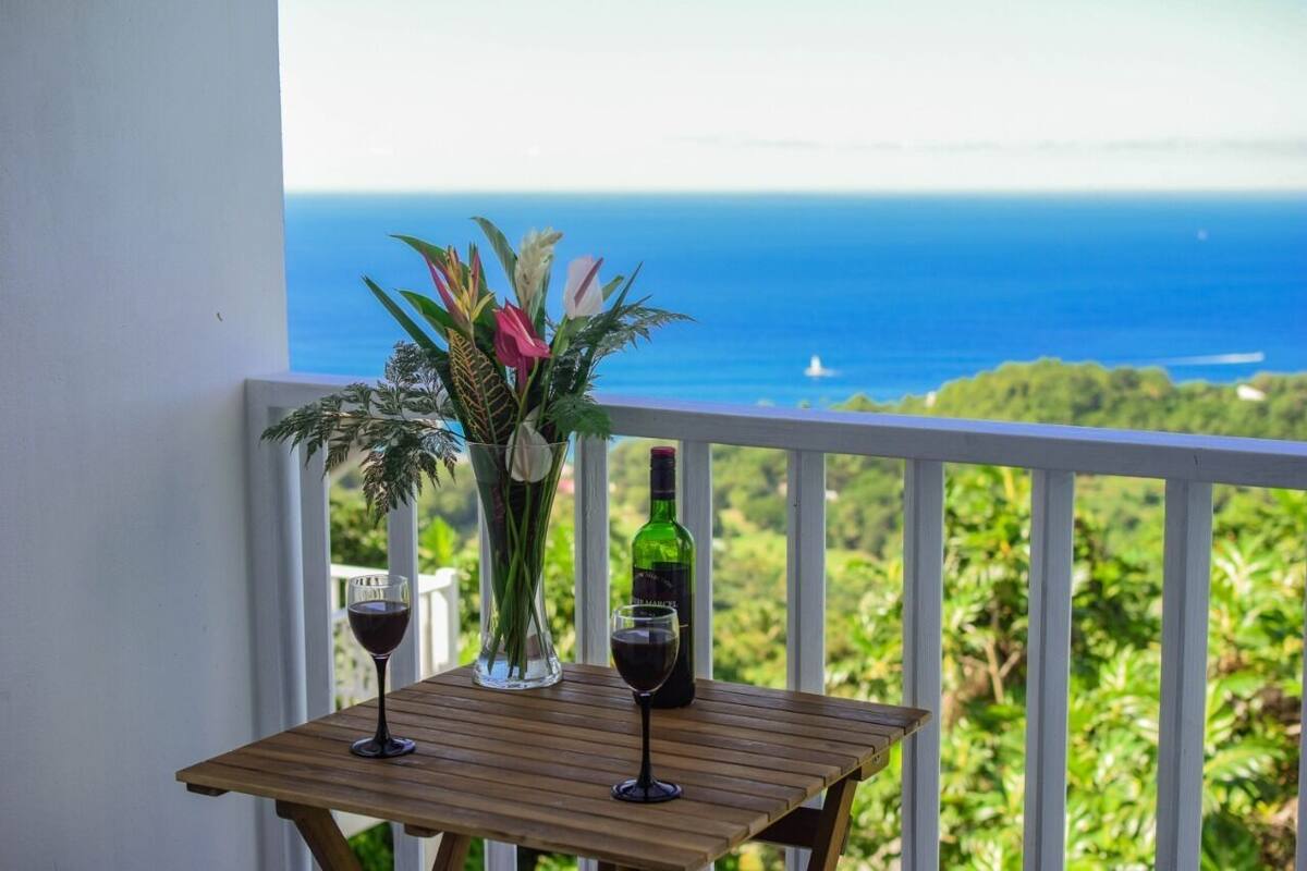 A wooden table with two wine glasses and a bottle of wine, a vase with flowers, and a view of the ocean.