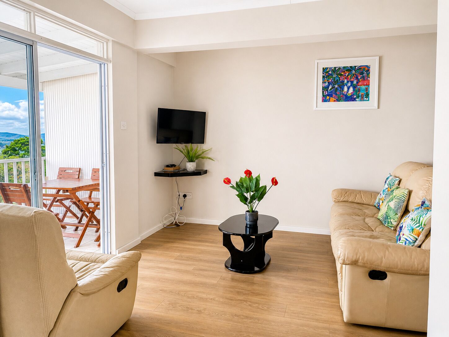 A living room with a sofa, a table with a plant, a TV, and a balcony with a table and chairs.