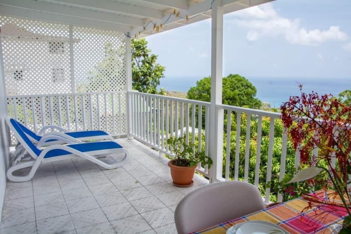 A balcony with two blue lounge chairs, a table with a colorful tablecloth, and a potted plant.