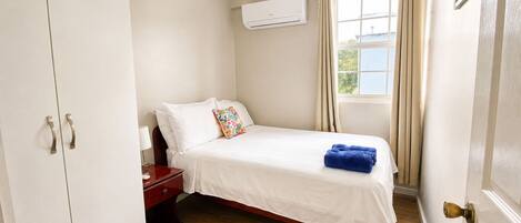 A bedroom with a bed, a nightstand, a wardrobe, an air conditioning unit, a window with curtains, and a view of the outdoors.