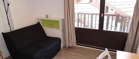 A room with a black sofa, a small table, a chair, a shelf with a candle, and a view of a wooden deck outside.