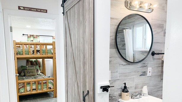 A bedroom with a bunk bed, a bathroom with a round mirror, and a sign above the door. - Whispering Pines by Cottonwood Lodging (Brighton)
