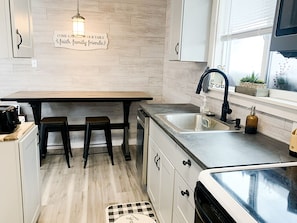 A modern kitchen with a wooden table, two stools, a sink, and a window with plants.