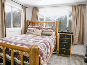 A bedroom with a wooden bed, patterned bedding, a nightstand with drawers, and a window with a mountain view.