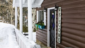 A wooden cabin with a porch covered by a white roof, a welcome sign, and a door. - Whispering Pines by Cottonwood Lodging (Brighton)
