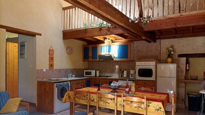 A cozy kitchen with wooden cabinets, a dining table with chairs, a washing machine, and a small dining area with a sofa and cushions. - Hyper-Gite: very large barn converted into gites (Corpe)