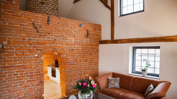 A cozy living room with exposed brick walls, a wooden ceiling, a brown leather sofa, and a window with a view.