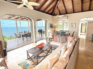 A spacious living area with a large sofa, a coffee table, and a dining table. There are multiple windows providing views of the outdoors.