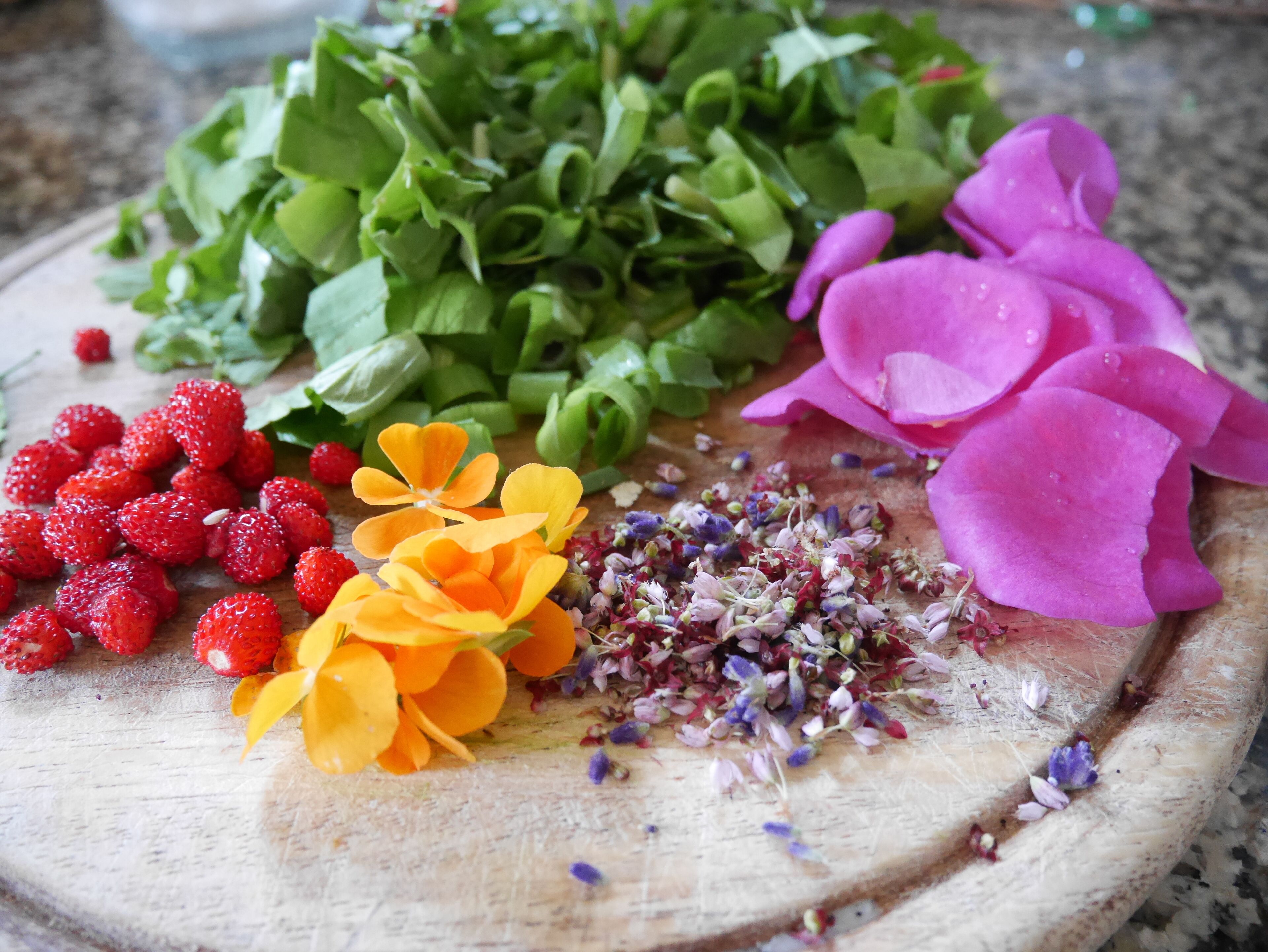 Un tagliere di legno con diversi ingredienti freschi, tra cui fragole, foglie verdi, fiori gialli, fiori viola e erbe essiccate.