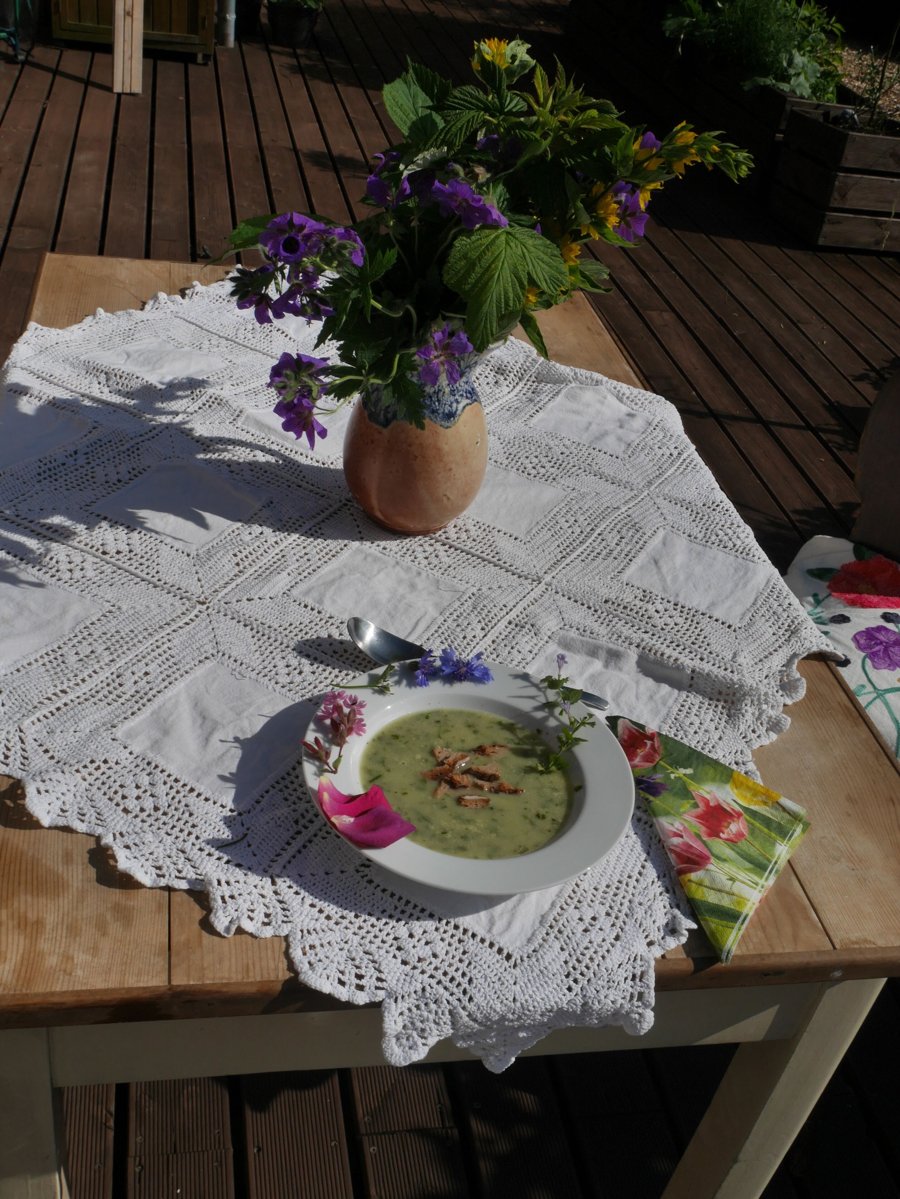 Ein Tisch mit weißer Spitzenleinentafel, eine Schüssel mit grüner Suppe, garniert mit Kräutern und Croûtons, ein Löffel und eine Tischservette mit Blumenmuster.