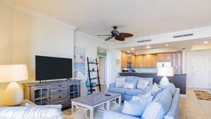 Una sala de estar con televisor, sofá y mesa de centro. Se observa una cocina al fondo con armarios y electrodomésticos.