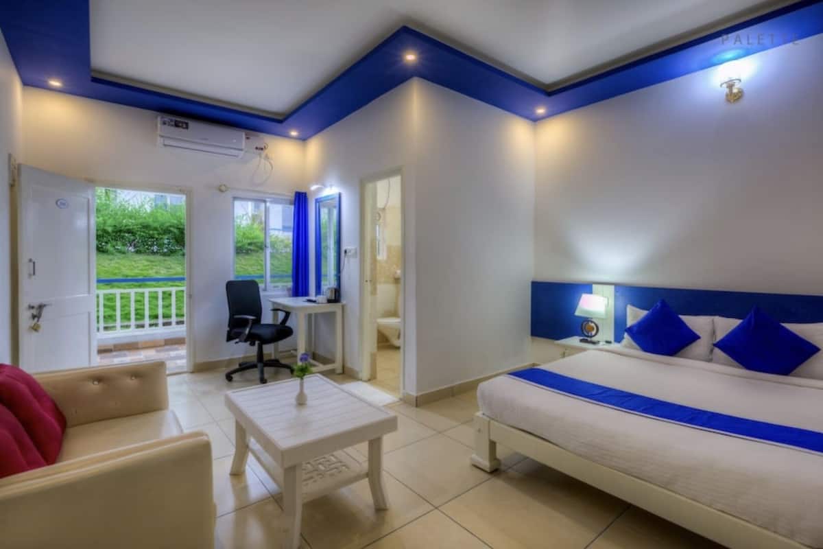 Quarto de hotel com uma cama, uma escrivaninha, uma cadeira e uma mesinha. Há uma janela com vista para o verde lá fora.