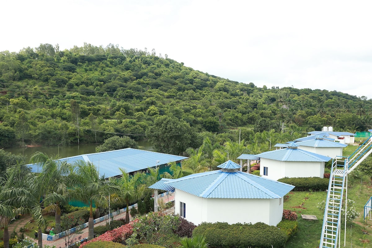 Resort com vários edifícios, cada um com telhado azul, rodeado por vegetação exuberante e uma colina ao fundo.