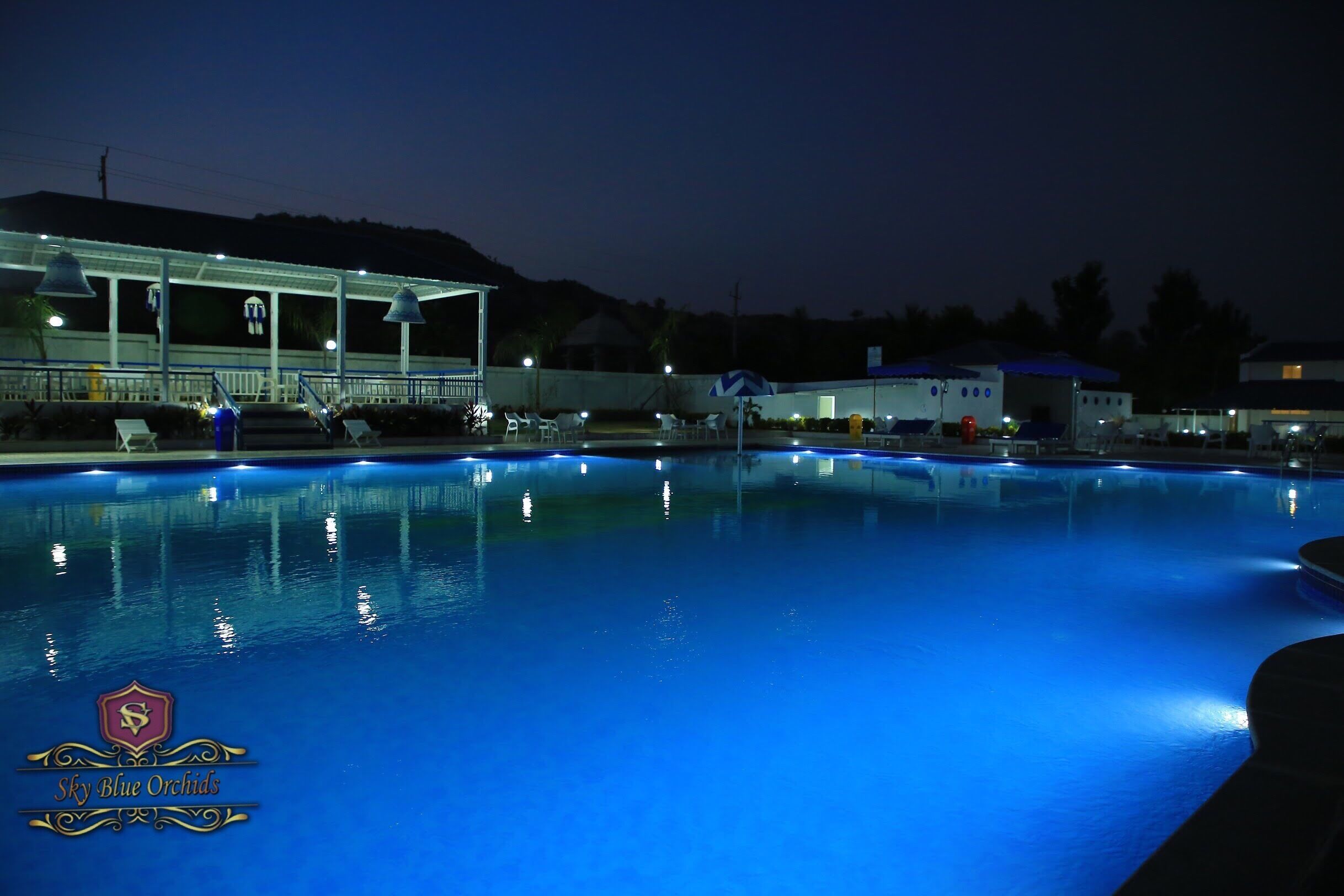 Un área de piscina iluminada de noche, con zona de estar cubierta y sillones reclinables.