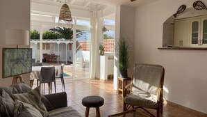 Ein geräumiges Wohnzimmer mit einer Couch, einem Sessel und einem Glastisch. Es gibt einen Deckenventilator, eine Hängeleuchte und einen Blick auf die Terrasse mit einem Esstisch und Stühlen.