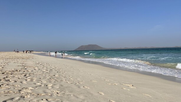 Ein weiter Sandstrand mit Fußspuren, die zum Wasser führen, ein klarer Himmel und ein Berg in der Ferne.