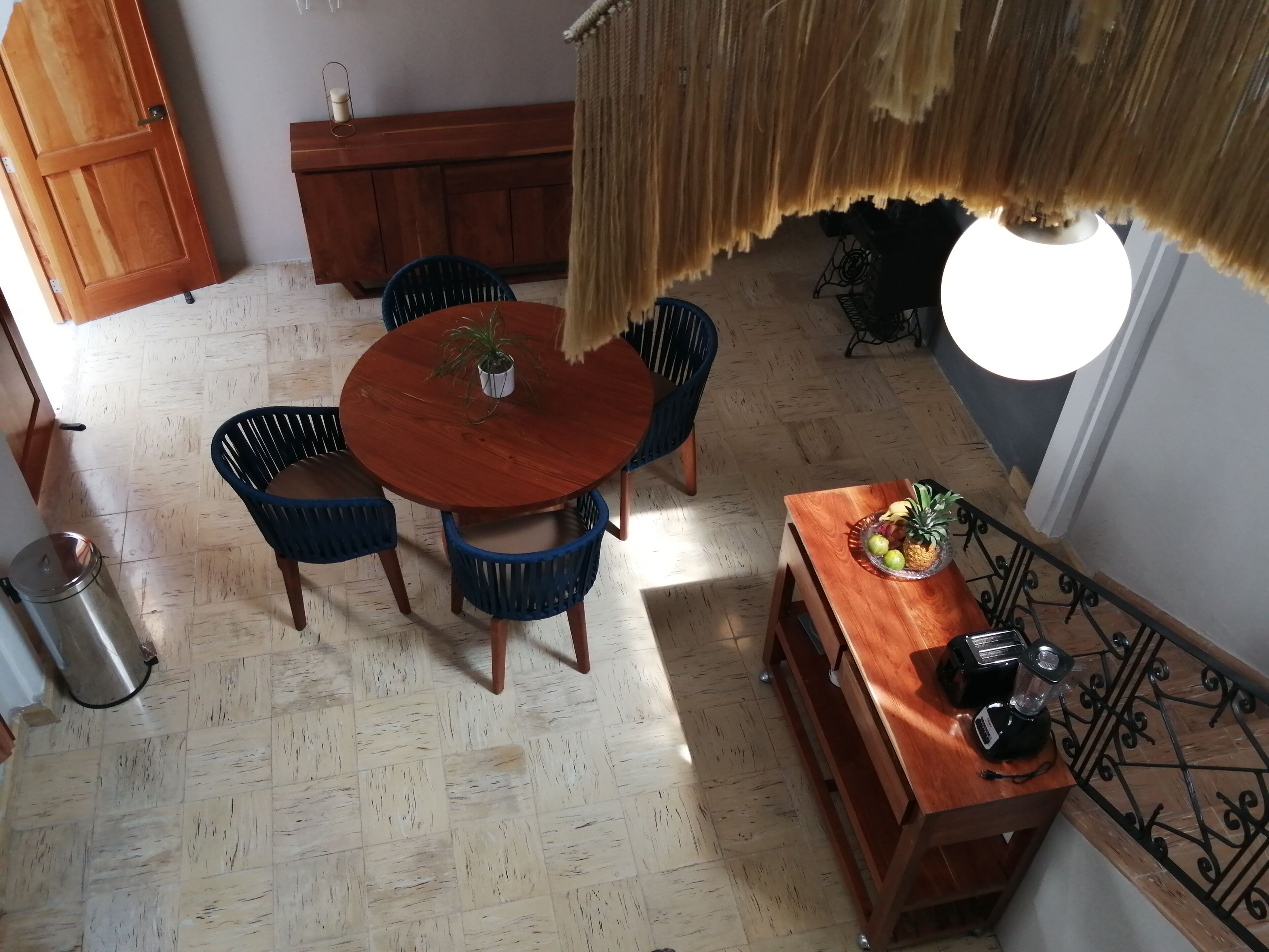 A dining area with a round wooden table, blue chairs, a potted plant, and a fruit bowl on a wooden side table.